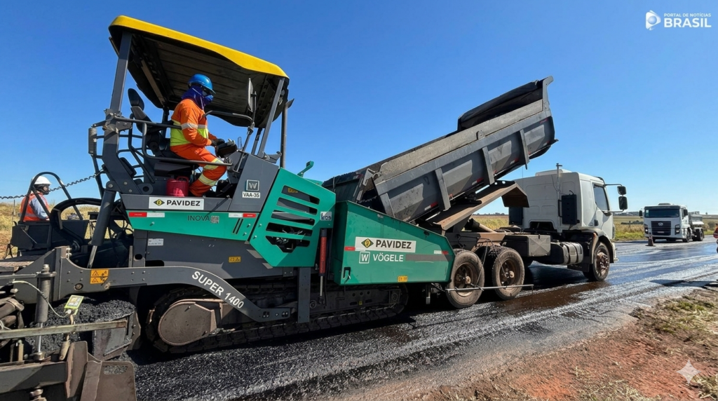 Máquina pavimentadora verde da Vögele trabalhando nas obras de duplicação da rodovia BR-163, com caminhão caçamba basculante ao fundo descarregando asfalto sob céu azul.