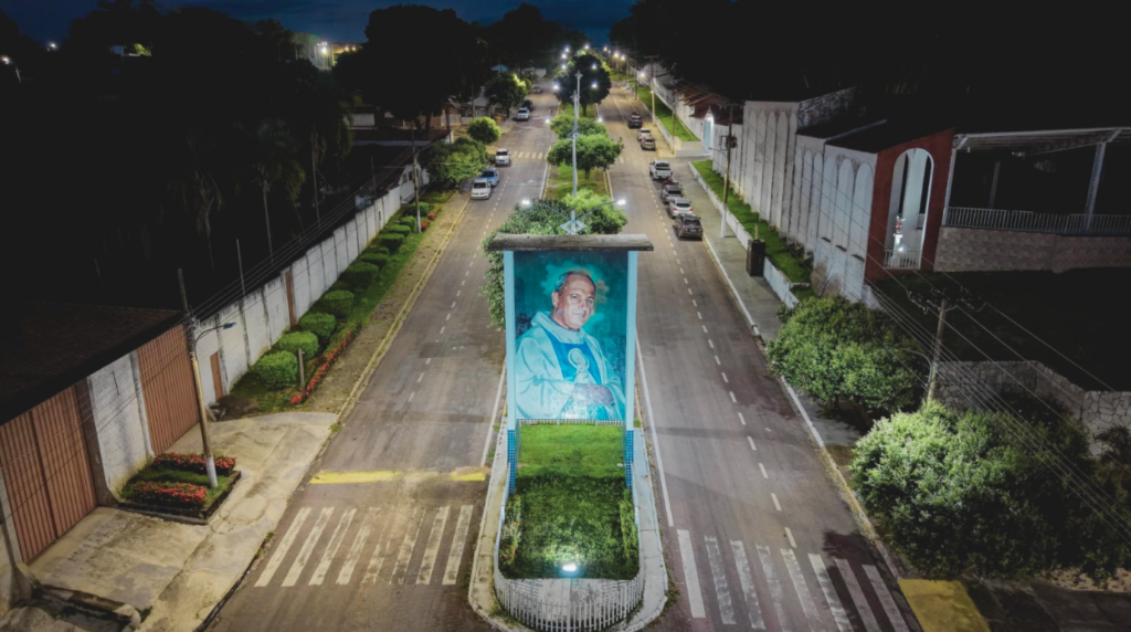 Imagem aérea noturna com iluminação otimizada mostrando o monumento em homenagem ao Padre Nazareno Lanciotti no canteiro central de uma avenida em Jauru, Mato Grosso, preparado para as celebrações de sua beatificação.