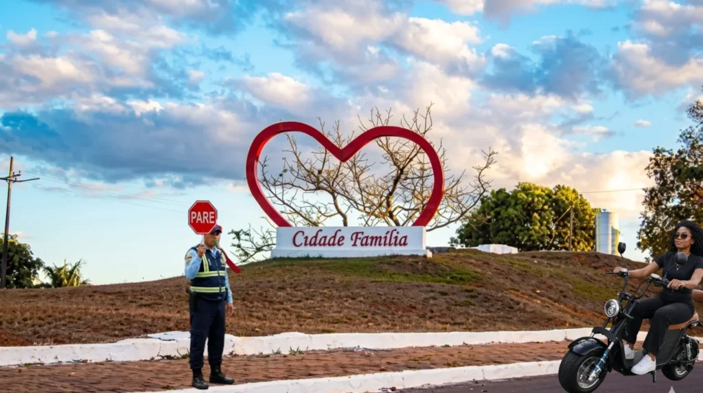 Criação digital IA. Monumento de coração da praça central de Colíder, em alvenaria e metal em forma de coração com os dizeres: Cidade Família. Guarda de trânsito com placa pare nas mãos e mulher conduzindo motocicleta elétrica.